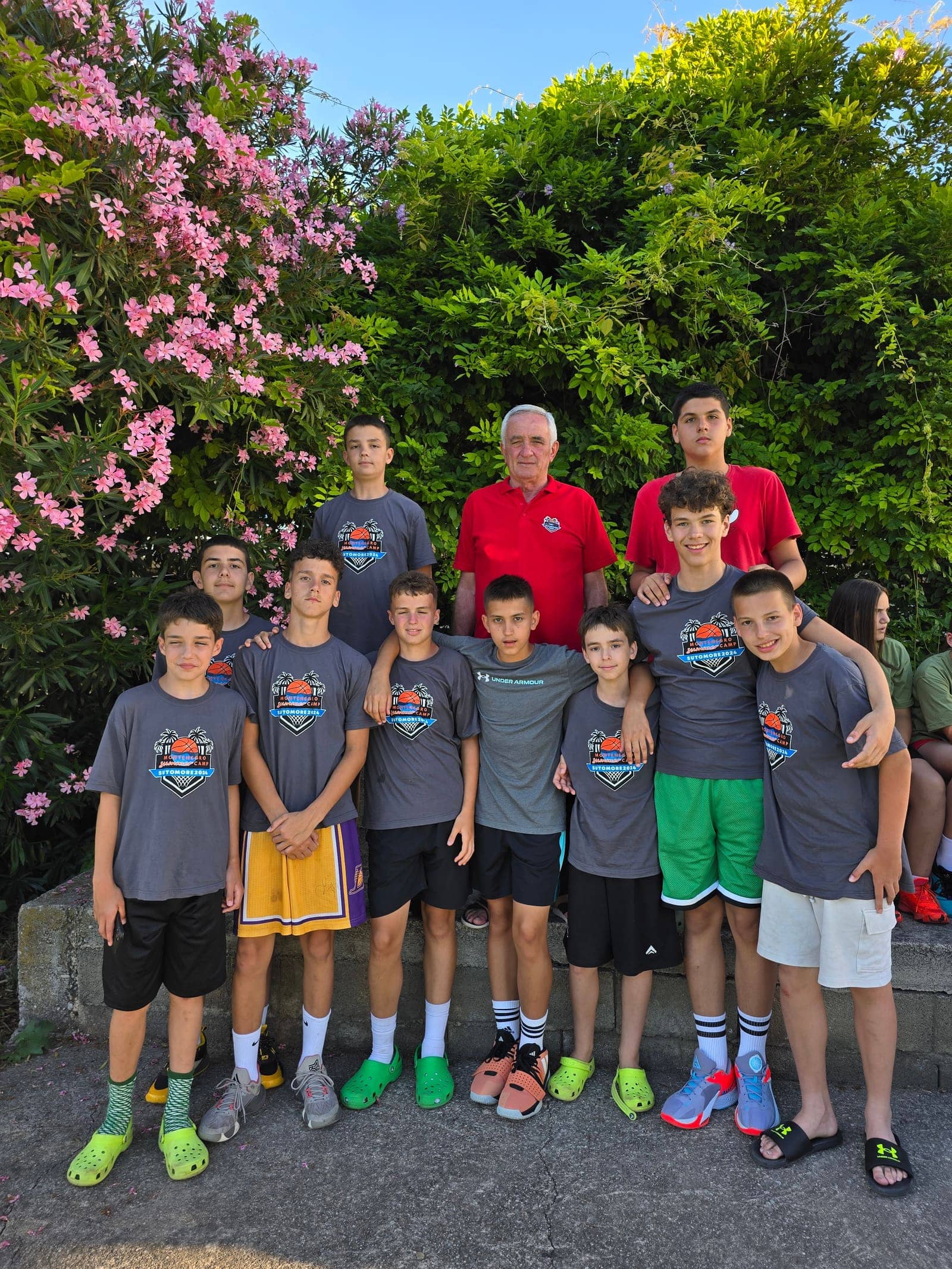 A coach and a group of boys in camp shirts in front of flowering oleander