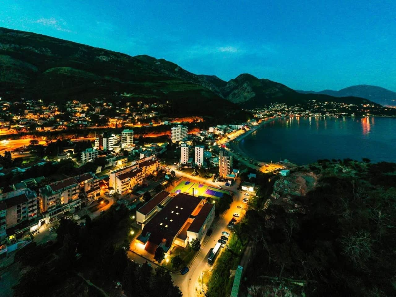 Sutomore from the air at dusk — the bay, town lights and the lit basketball courts beside the accommodation